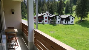 Un balcon avec un banc en bois et une table, donnant sur un espace verdoyant où l’on aperçoit plusieurs bâtiments blancs au loin.
