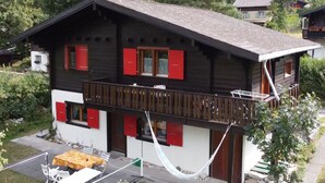 Een chalet met twee verdiepingen en een balkon, omringd door weelderige begroeiing en bergen.
