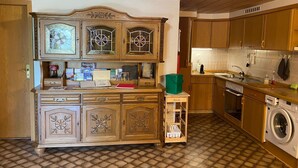 Een keuken met houten kastjes, een inbouwfornuis, een wasmachine en een klein tafeltje met een groene emmer.