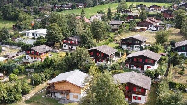Een groep traditionele chalets met schuine daken, omgeven door weelderige begroeiing en bergen op de achtergrond.