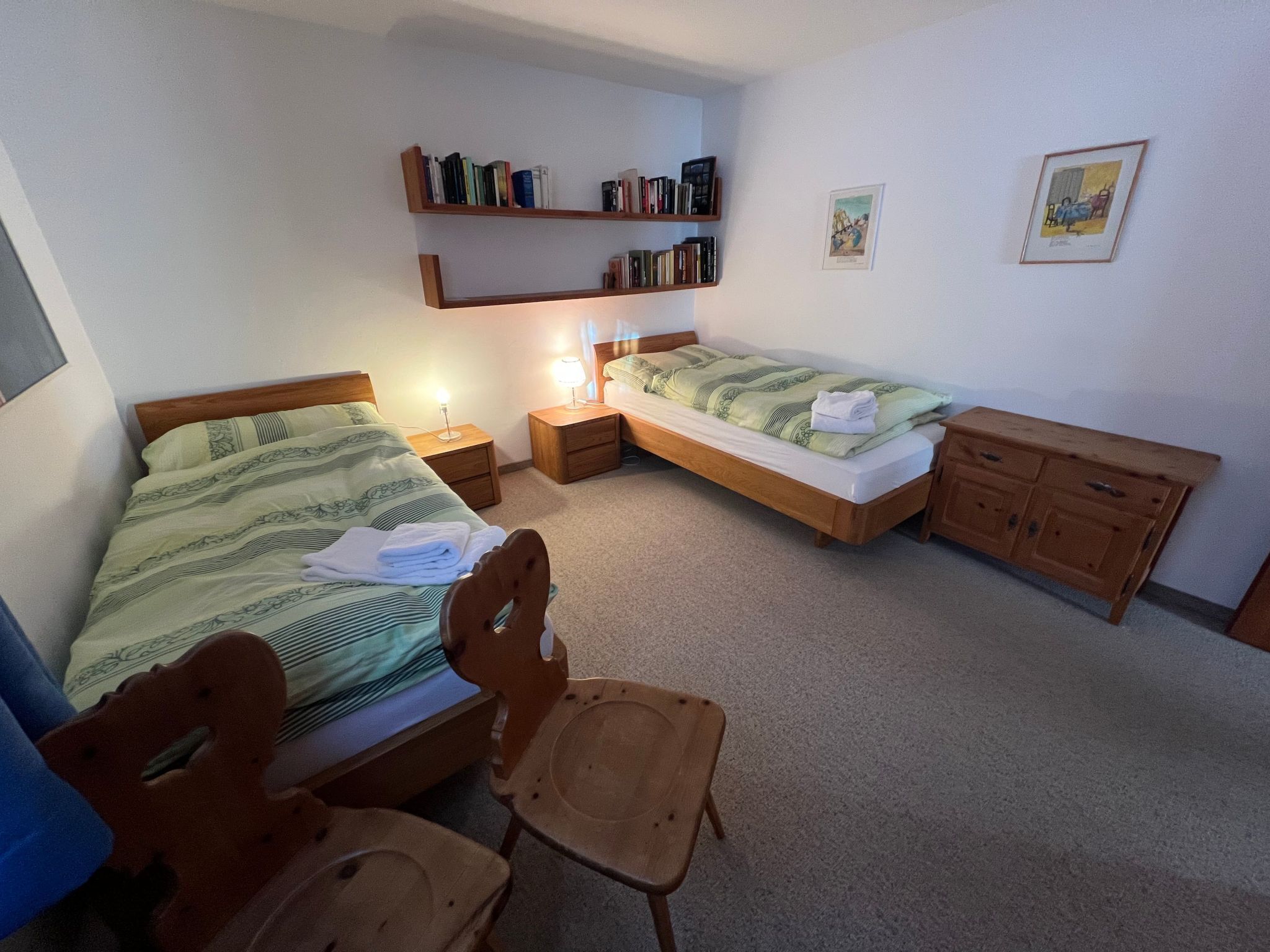 Une chambre avec deux lits, une commode en bois, une chaise, une table de chevet avec une lampe et une bibliothèque.