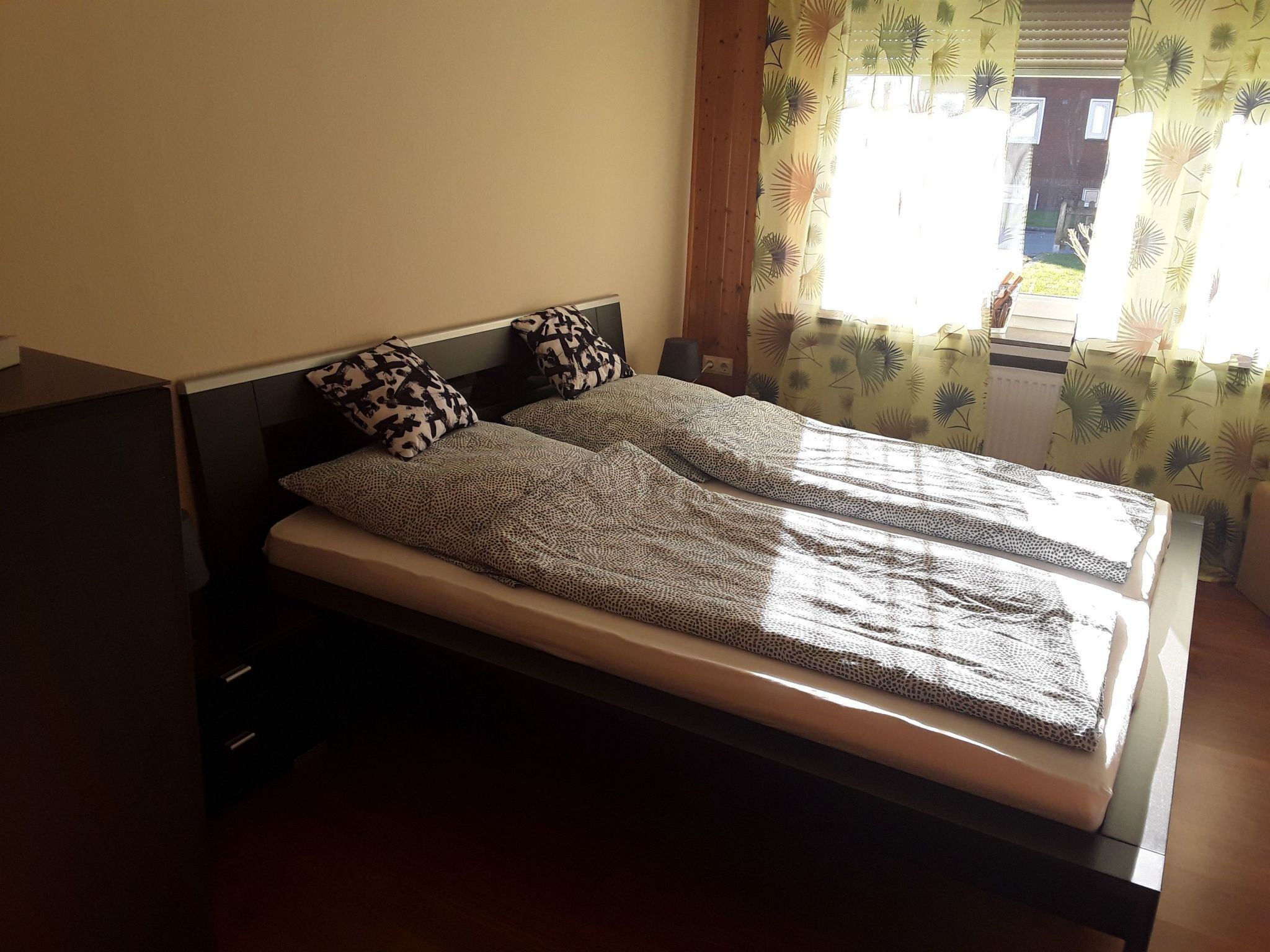 Ein Bett mit gemusterten Bettbezügen und Kissen, ein hölzernes Kopfende und ein Fenster mit Vorhängen.