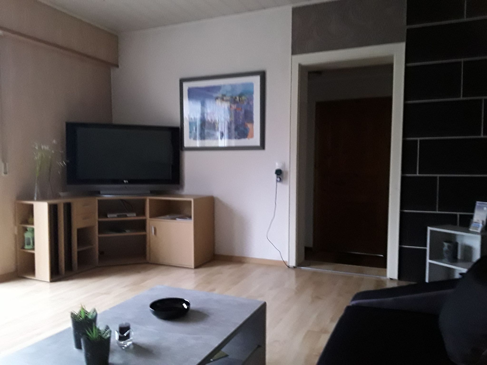A living room with a flat-screen TV on a wooden stand, a framed picture on the wall, and a doorway leading to another room.