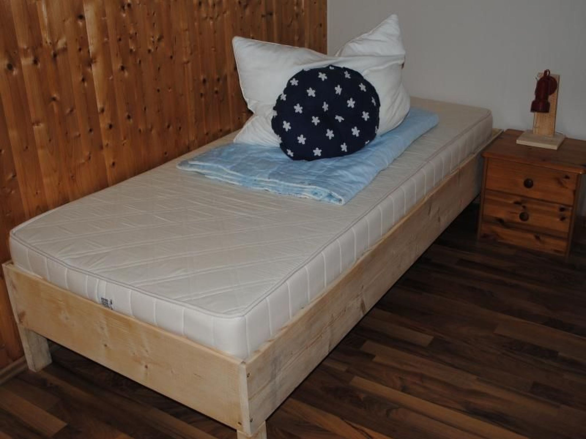 A single bed with a white mattress, a blue blanket, and a pillow with white stars. There is a wooden nightstand with a small wooden figure on it. The room has wooden flooring and a wooden headboard.