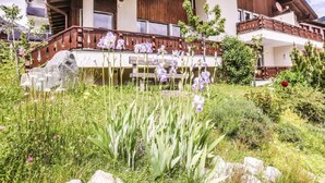 Un bâtiment de style chalet, doté d’un balcon en bois, entouré d’un jardin où poussent différentes plantes et fleurs.