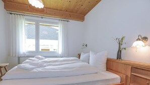Une chambre à coucher avec un lit en bois, du linge de lit blanc, une fenêtre avec des rideaux, une table de chevet et un luminaire fixé au mur.