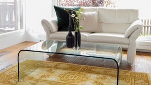 Un salon avec une table basse en verre, un tapis à motifs, un canapé blanc et une fenêtre donnant sur des arbres.