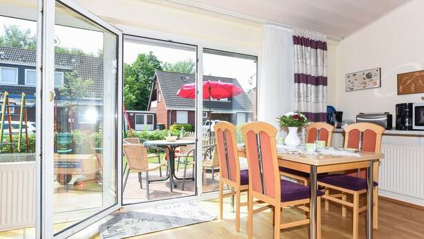 A dining area with a table and chairs, a kitchen area with appliances, and a sliding glass door leading to an outdoor patio. - Strandnahes Ferienhaus mit Kinderspielplatz by Interhome (Bensersiel)