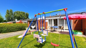 A playground with swings and a slide, a patio area with tables and chairs, and a red umbrella. - Strandnahes Ferienhaus mit Kinderspielplatz by Interhome (Bensersiel)