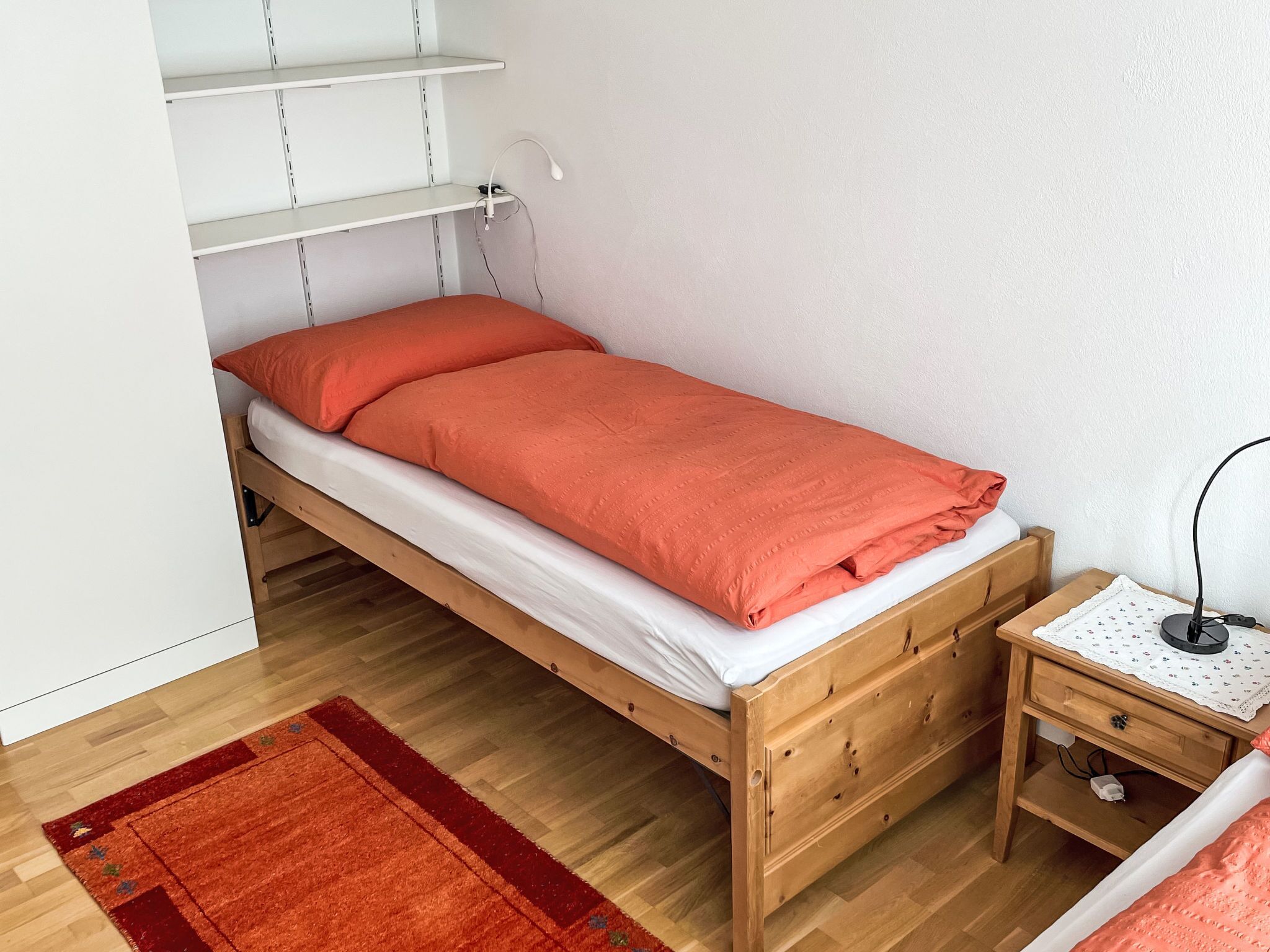 Un lit simple avec un cadre en bois, une couverture rouge, une petite table de chevet avec une lampe et un tapis rouge sur un plancher en bois.