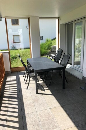 A covered patio area with a table and chairs, large sliding glass doors, and a view of a grassy yard. - Fewo mit Bergblick by Interhome (Savognin)