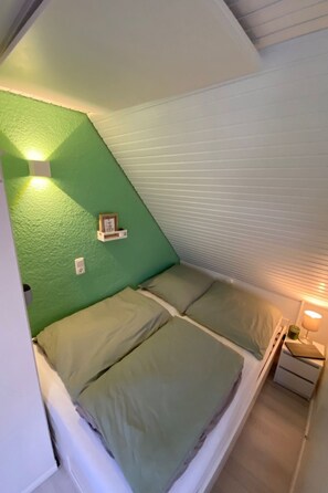 Quarto pequeno e acolhedor com teto inclinado, parede verde e cama com roupa de cama verde. Há uma pequena prateleira com um porta-retrato e uma luminária na mesinha de cabeceira.