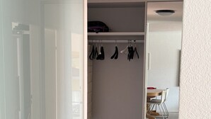 Un dressing accessible sans porte, doté d’un espace pour suspendre les vêtements et d’étagères, d’un plancher en bois et d’une vue sur une autre pièce où se trouvent une chaise et une table.