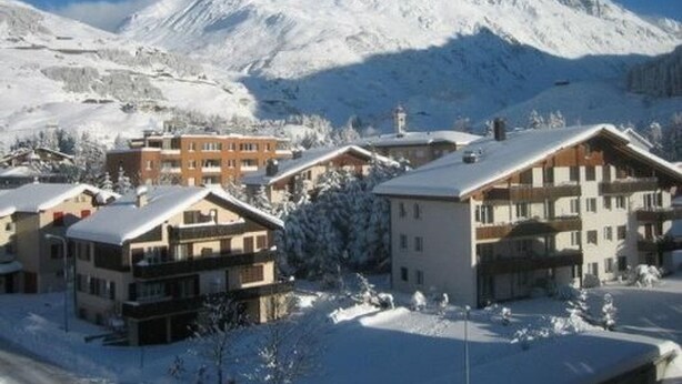 Une station de montagne enneigée, avec des bâtiments de style chalet, un ciel dégagé et une piste de ski en arrière-plan.