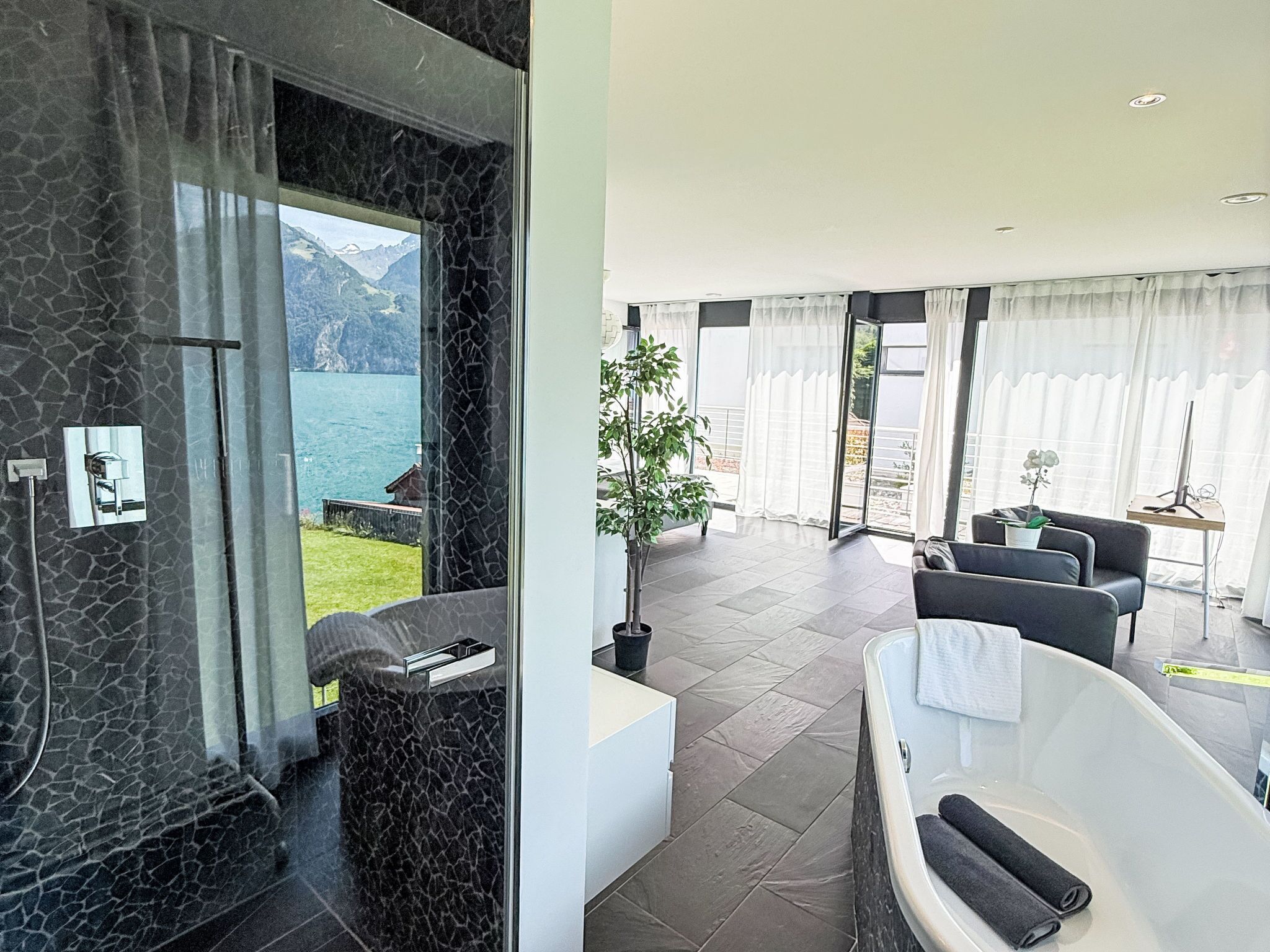 Un baño moderno con bañera, zona de ducha y puerta de cristal que da a un balcón con vistas a un lago y montañas.