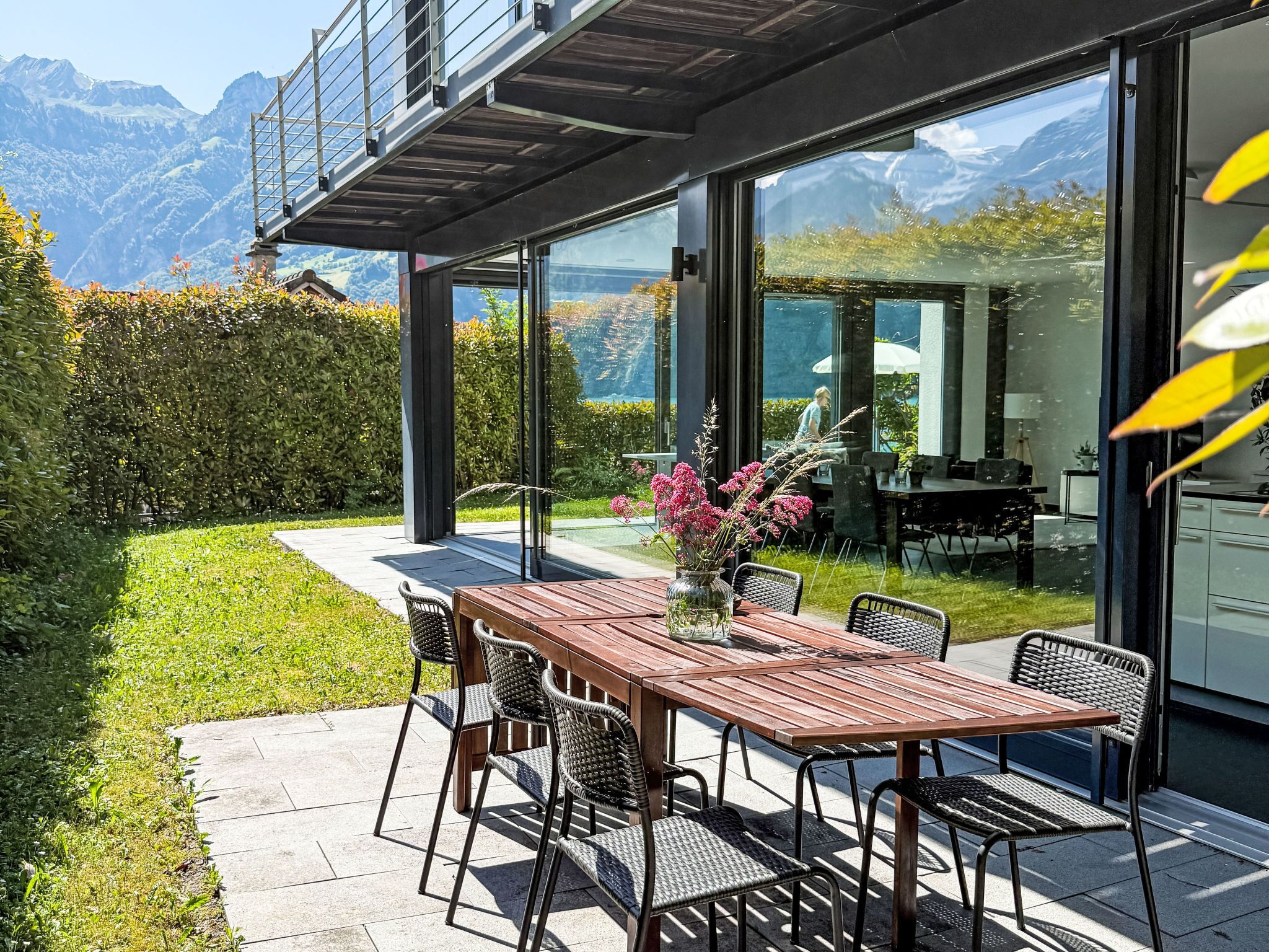 A modern outdoor dining area with a wooden table and chairs, a glass-enclosed kitchen, and a view of mountains.