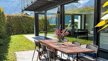 A modern outdoor dining area with a wooden table and chairs, a glass-enclosed kitchen, and a view of mountains.