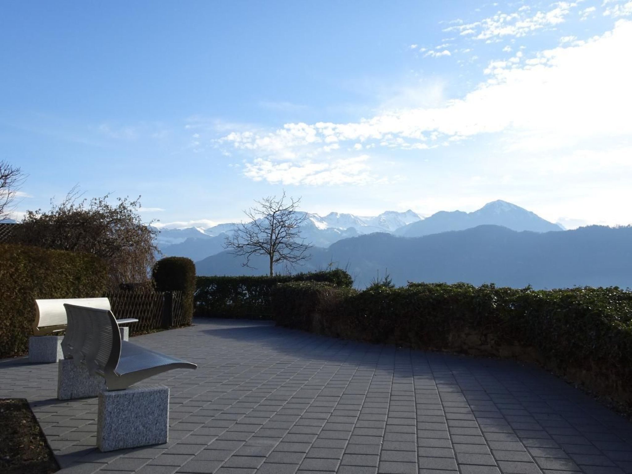 Ein gepflasterter Außenbereich mit einer Bank und einer Skulptur, mit Blick auf ein Gebirge.