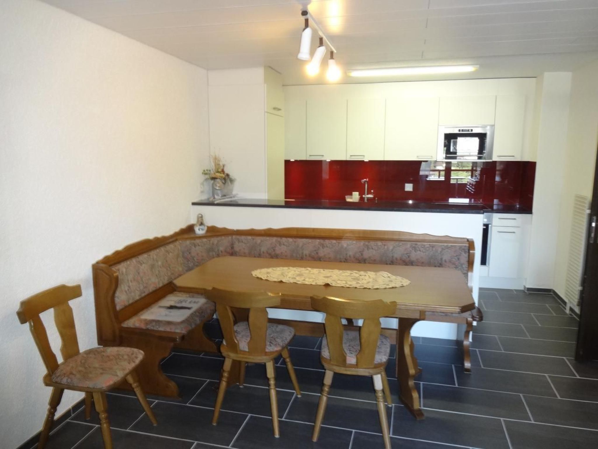 Un coin repas avec une table et des chaises en bois, une cuisine en arrière-plan avec des meubles blancs et un pare-feu rouge, et un sol carrelé.
