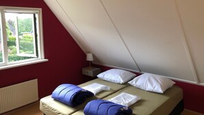 Una habitación pequeña con cama individual, techo inclinado, ventana con vistas a follaje, mesita de noche con lámpara y radiador.