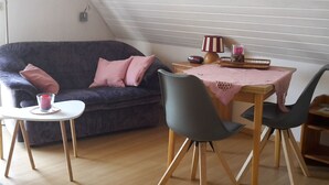 Una sala de estar acogedora con un sof á azul oscuro, dos sillas grises, una mesita blanca y una mesa de comedor de madera con mantel rosa.
