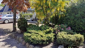 Un jardín ajardinado con arbustos variados y un árbol con flores amarillas, un sendero pavimentado y un coche estacionado.