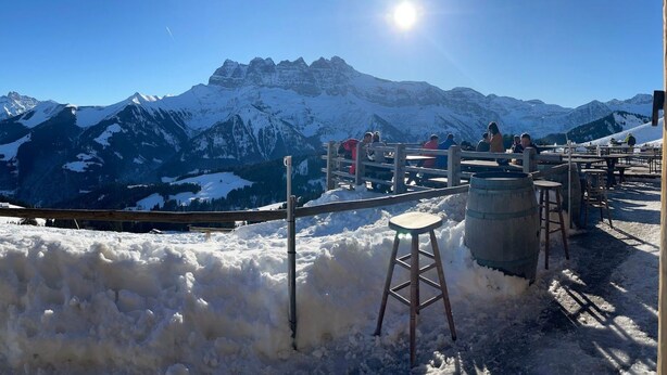 Un área de estar al aire libre cubierta de nieve con mesas y sillas de madera, con vistas a una cadena montañosa bajo un cielo azul despejado.