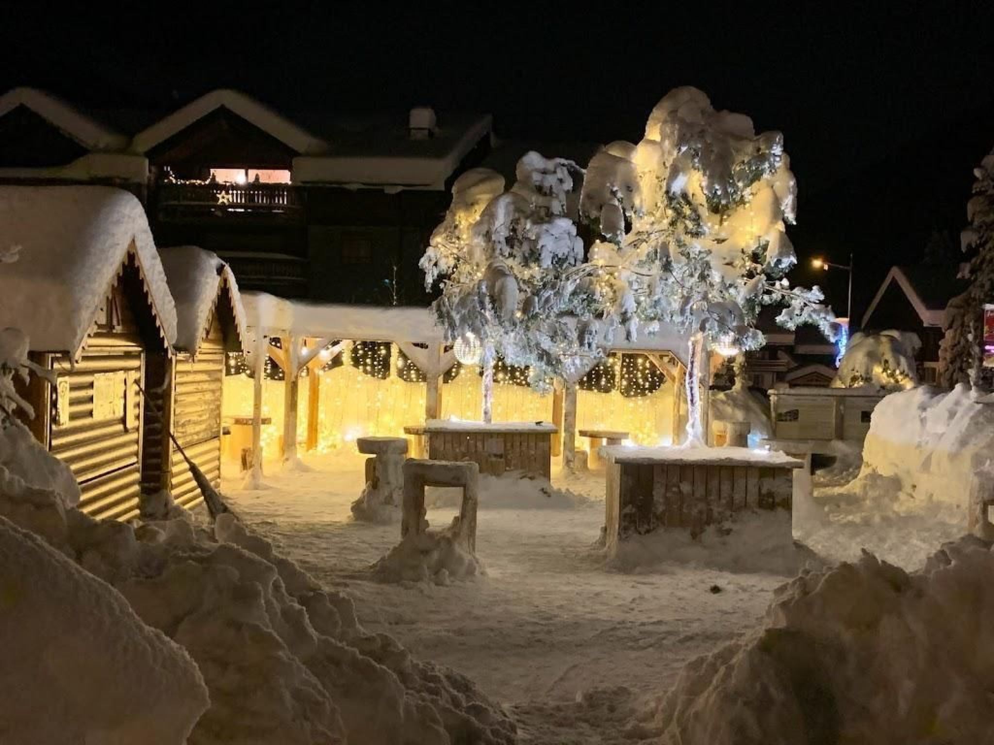 A snow-covered outdoor area with benches and a wooden structure, likely a lodge or cabin, with lights illuminating the scene.