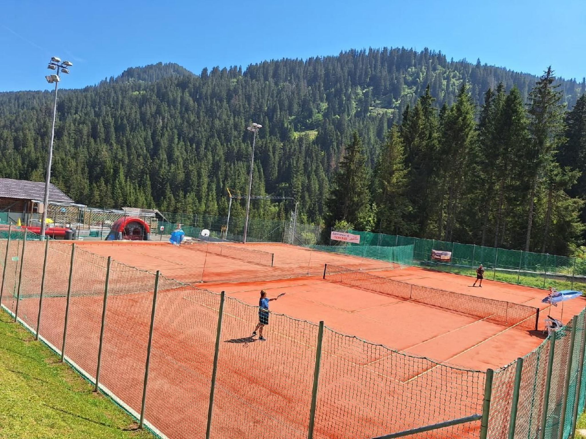 An outdoor tennis court with a net, surrounded by a fence and a forested mountain in the background.