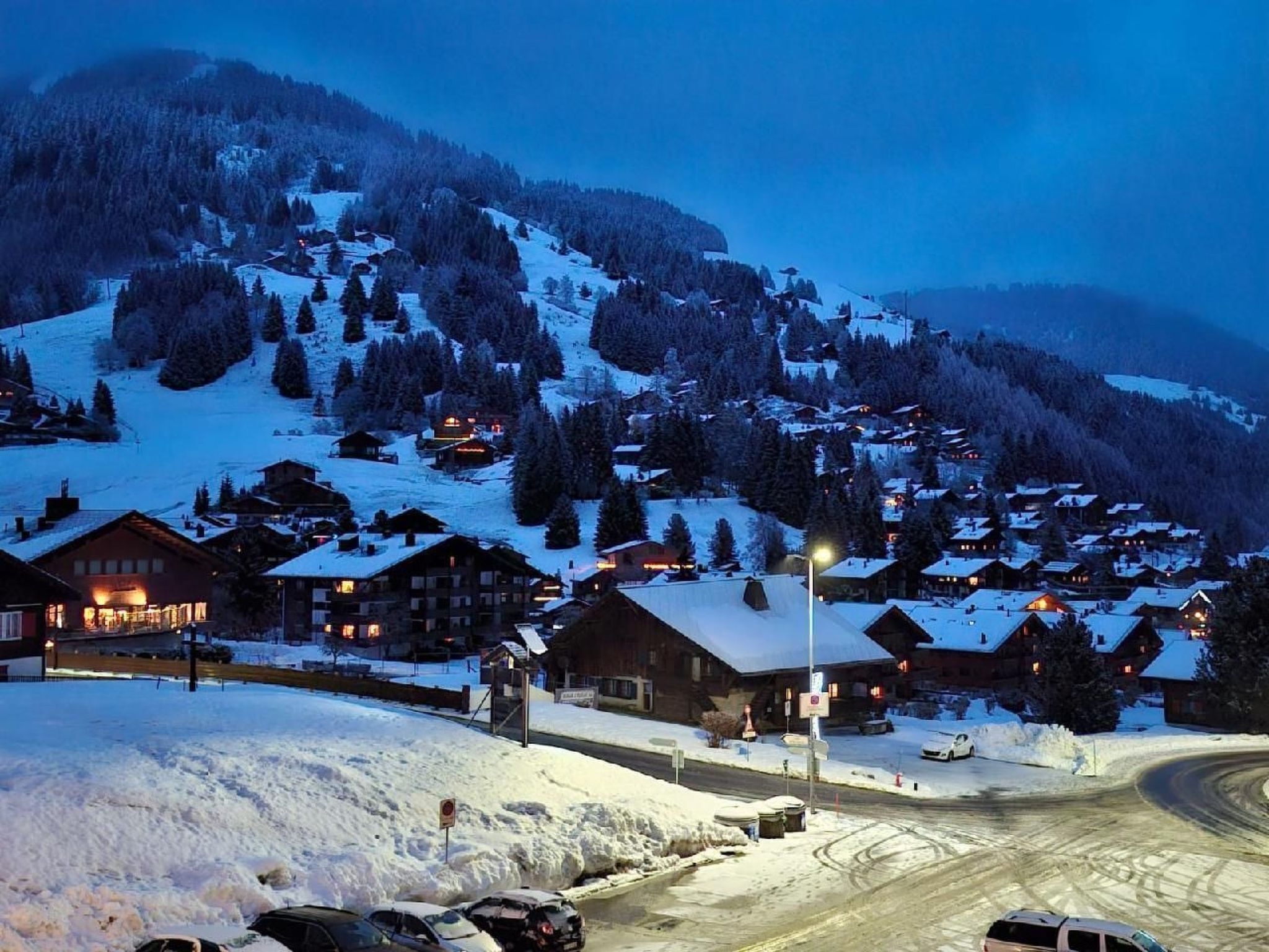 Un village de montagne enneigé, au crépuscule, avec des bâtiments et une route.