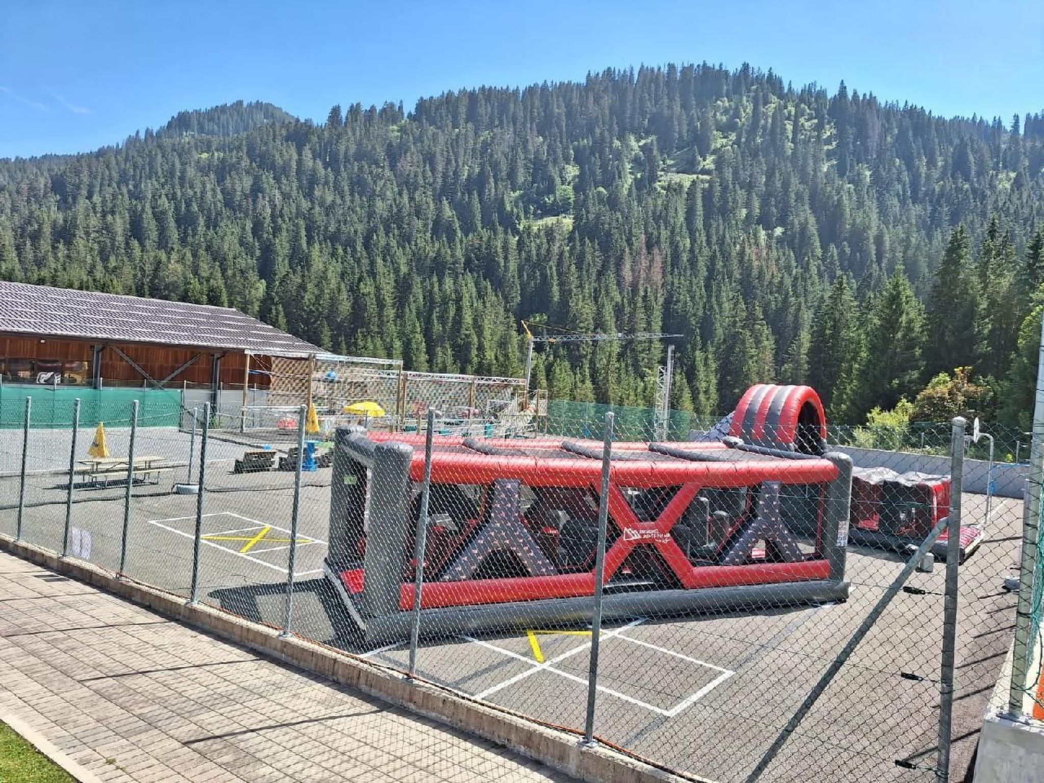 An outdoor trampoline area with a fenced enclosure, a building in the background, and a forested hill.
