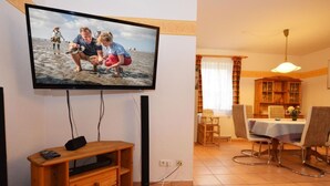 Ein Wohnzimmer mit Flachbildfernseher an der Wand, einer Holz-Unterhaltungsecke, einem Essbereich mit Tisch und Stühlen sowie einer Küchenzeile mit Schrank.