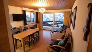 Ein gemütliches Zimmer mit Holzbalkendecke, einem Esstisch mit Stühlen, einer Couch mit Kissen, einem an der Wand befestigten Fernseher und einem großen Fenster mit Blick auf verschneite Berge.