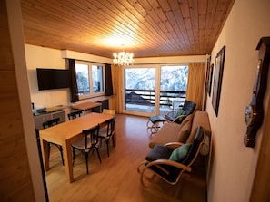 A cozy room with a wooden ceiling, a dining table with chairs, a sofa with cushions, a TV mounted on the wall, and a large window with a view of snowy mountains. - 3 Zi FeWo App 23. Le Miroi an Skipiste by Interhome (Les Crosets)