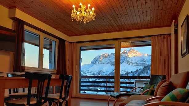 Ein Zimmer mit Holzbalkendecke, einem Chandelier, einem Esstisch mit Stühlen, einer Couch mit Kissen und großen Fenstern mit Bergblick.