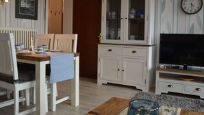 Uma área de refeições com uma mesa posta para dois, um armário branco com portas de vidro, uma TV de tela plana sobre um suporte de madeira e um relógio de parede.