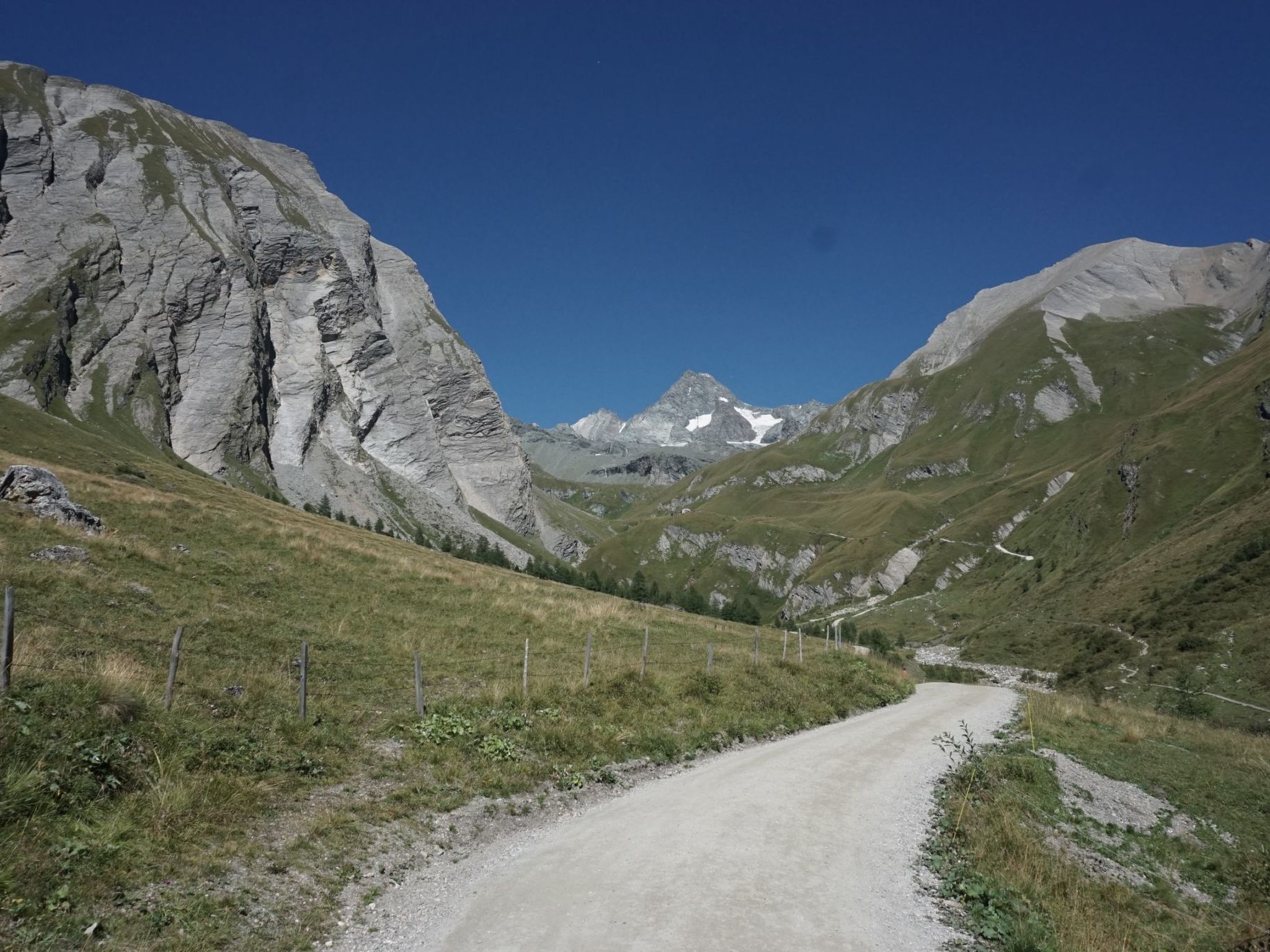 Una strada di montagna con cielo sereno, circondata da montagne rocciose e vegetazione rada.