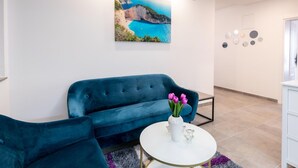 Una habitación moderna con un sofá azul, un jarrón blanco con flores y un cuadro de un paisaje costero.