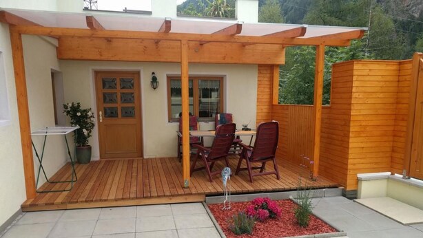 Ein überdachter Patio-Bereich mit einer Holzhütte, einem Esstisch mit Stühlen, einem Blumentopf und einem kleinen Blumenbeet.