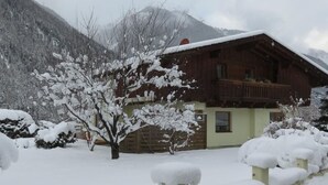 Eine Holzhütte mit Balkon, umgeben von schneebedeckten Bäumen und Bergen im Hintergrund.