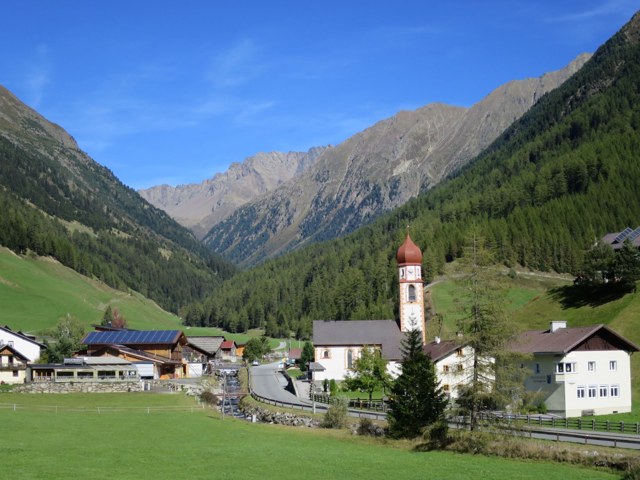 A mountain village with a prominent church tower, houses with solar panels, and a clear view of the surrounding mountains.