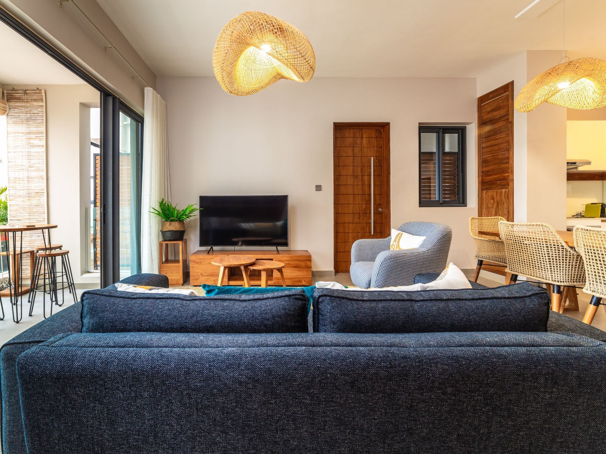 Uma sala de estar moderna com um sofá grande azul-escuro, uma televisão de tela plana, luminárias pendentes de vime e um espaço de jantar com cadeiras de vime.