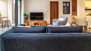 Ein modernes Wohnzimmer mit einem großen, dunkelblauen Sofa, einem Flachbildfernseher, Korbstuhl-Pendelleuchten und einem Essbereich mit Korbstühlen.