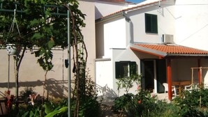 A two-story building with a white facade, red-tiled roof, and green shutters. There is a garden area with various plants and a pergola with climbing vines. - Gemütliche Ferienwohnung Emil by Interhome (Dragišina)