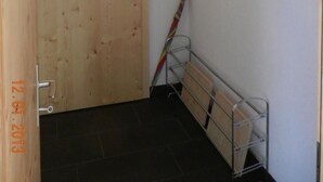 Ein Zimmer mit Holztür, einem Metallgestell mit Kleiderbügeln, einem roten hängenden Gegenstand und einem an der Wand angelehnten Besen.