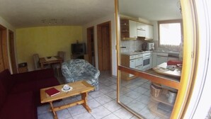 Ein Wohnzimmer mit Sofa, Tisch und Stuhl. Durch eine Glastür ist eine Küche zu sehen.