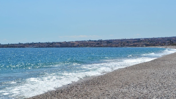 Une plage côtière au rivage de galets, avec une mer bleue et limpide, et une ville qui se devine au loin.
