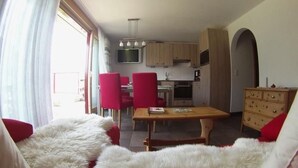 A cozy living room with a fur-covered sofa, a wooden dining table, and red chairs. A kitchen with wooden cabinets and a TV is visible in the background. - Alvetern Marmotta by Interhome (Scuol)