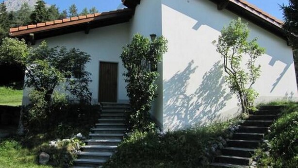 Edificio a due piani con facciata bianca e tetto marrone. Scale conducono all'ingresso, circondate da vegetazione.