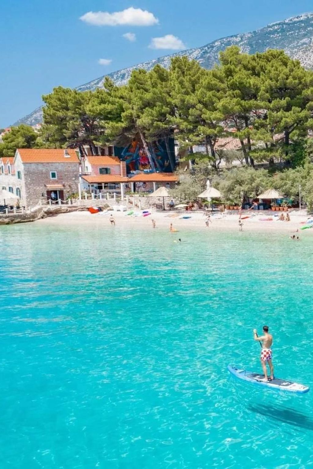 Een kustlandschap met helder turquoise water, een persoon op een paddleboard, en gebouwen met rode pannendaken op de achtergrond.